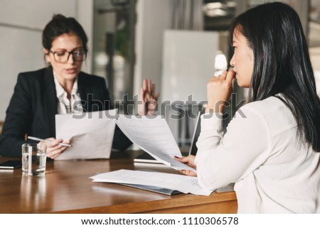 Image of nervous asian woman looking and talking to businesswoman while sitting at table in office during job interview - business, career and recruitment concept Royalty-Free Stock Photo #1110306578
