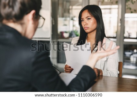 Image of serious asian woman looking and talking to businesswoman while sitting at table in office during job interview - business, career and recruitment concept Royalty-Free Stock Photo #1110306590