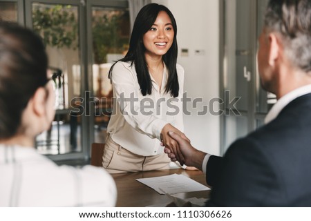 Business, career and placement concept - image from back of two employers sitting in office and shaking hand of young asian woman after successful negotiations or interview Royalty-Free Stock Photo #1110306626