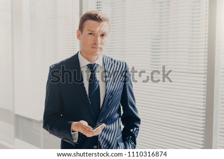Prosperous male director sends message and multimedia files via smart phone, synchronizes phone data and shares photos for project, poses in office, wears luxury expensive suit, looks seriously
