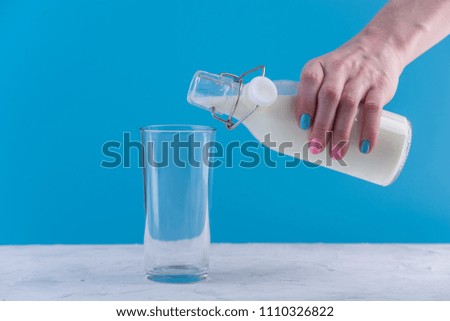 A woman's hand pours fresh milk from a glass bottle into a glass on a blue background. Colorful minimalism. The concept of healthy dairy products with calcium