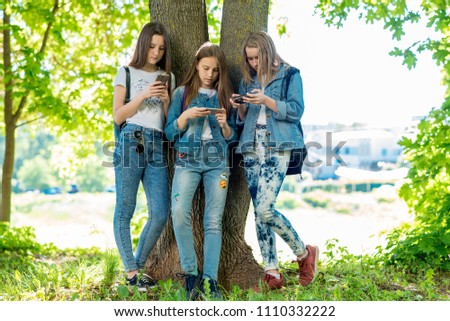 Three teenage girls stand by tree in summer park. In his hands holds smartphone and communicates social networks. Corresponds with parents concept of internet addiction. Friendly communication.