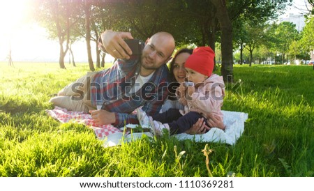 Happy family is laying on the grass and doing selfie with a baby at sunset in the park. Father and mother take pictures of themselves with the baby on the phone