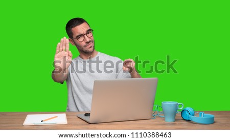 Serious young man with a stop gesture sitting at his desk - Green background