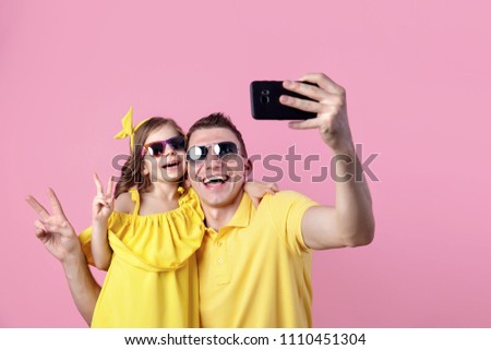 Portrait of happy family of two people in yellow. Daddy and little daughter doing selfie with smartphone on pink background. Horizontal color photography.
