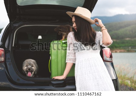 Happy young woman traveler sitting in hatchback car with dogs while parking near lake with mountain view and sunset. Female in white dress enjoys travel with pets in summer vacation in road trips.