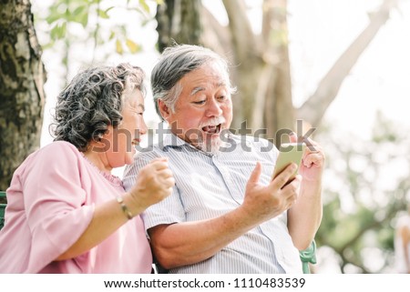 Happy senior Asian couple laughing and exciting with something in smartphone mobile while sitting in the park Royalty-Free Stock Photo #1110483539