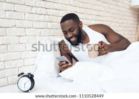 Black, awakened man drinks coffee in bed browsing web. Early morning. Pleasant awakening.