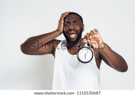 African American man holds head with alarm clock in hand, overslept his work. Isolated on white background. Studio portrait.