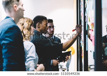 Pensive diverse group of young people dressed in formal wear writing notes on colorful stickers glued on wall during collaborative process in office.Multicultural students cooperating on common task Royalty-Free Stock Photo #1110549236