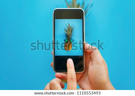 fruit photo. hands holding phone and taking photo of pineapple in sunglasses on blue paper, trendy flat lay. stylish food photography. creative phone image