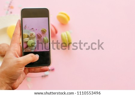 hand holding phone and taking photo of tasty colorful macarons in plate on trendy pastel pink paper flat lay. space for text. modern food photography concept. instagram photo workshop