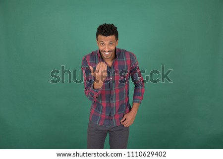 Handsome young man wearing a casual outfit, and he is calling someone to come, on his face big smile, standing on a green background.