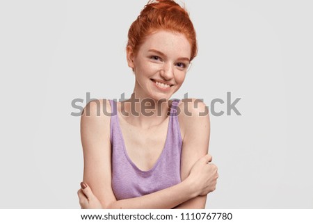 Studio shot of glad shy beautiful female with freckled, keeps hands crossed, has positive smile with white teeth, poses against white background. Red haired youngster shows her natural beauty