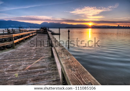 Pier moving out to the water while sun star rises over the mountain and Vancouver skyline can be seen to the right of the photo.