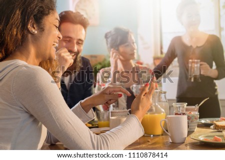 A group of multi-ethnic friends gathered around a table in the kitchen for breakfast. Two friends look at a picture on a phone