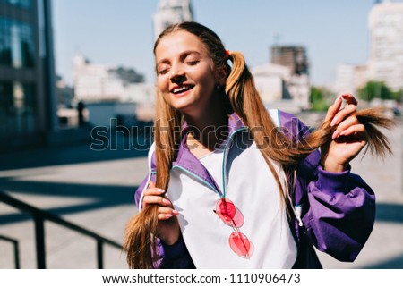 Outdoor lifestyle portrait of young beautiful fashionable girl posing at camera with lovely smile and closed eyes in morning sunshine. Female fashion concept, true happy emotions, summer sunny days