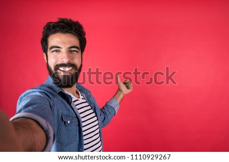 Handsome young man wearing a casual outfit taking a selfie by his phone, raising his thumb up, standing on a red background.