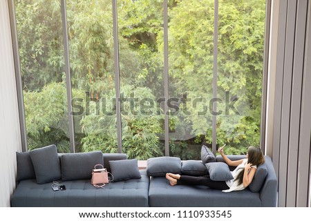 Asian woman laying sitting and taking selfie on sofa near big glass windows, relaxing alone in house or apartment with green forest in background, concept for people who living in modern lifestyle. Royalty-Free Stock Photo #1110933545