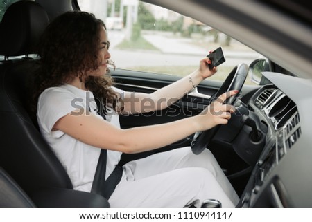 Smiling young woman taking selfie picture with smart phone camera driving a car.