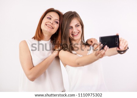 Two happy girls taking a selfie in studio on a white wall
