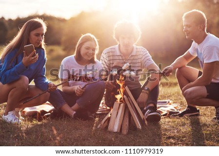 Photo of happy classmates or friends roast marshmallows over campfire, sit on ground, have picnic together during summer day, nice sunny weather, have pleasant talk. Togetherness and lifestyle concept