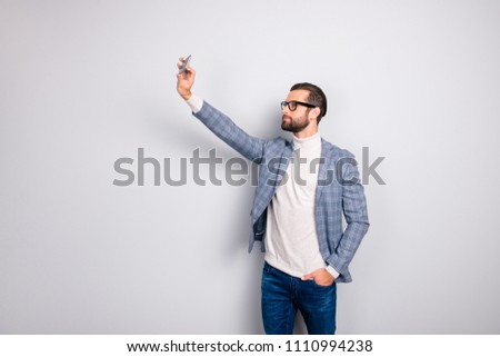 Virile, harsh, education, attractive man in jacket shooting self picture on smart phone front camera, holding hand in pocket of pants, standing over gray background