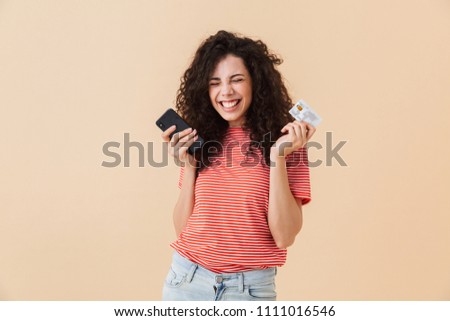 Photo of cute pretty young curly woman isolated over beige background using mobile phone holding credit card.