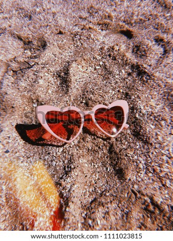 Pink heart sun glasses in the picture. There is a brown sand at the background.