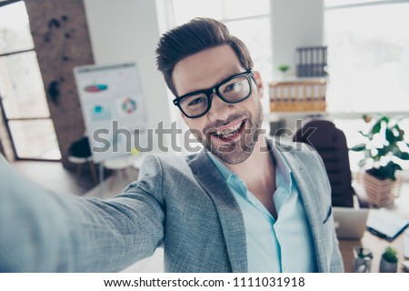 Self portrait of joyful funny man in shirt and jacket making selfie on smart phone with hand, standing in workplace