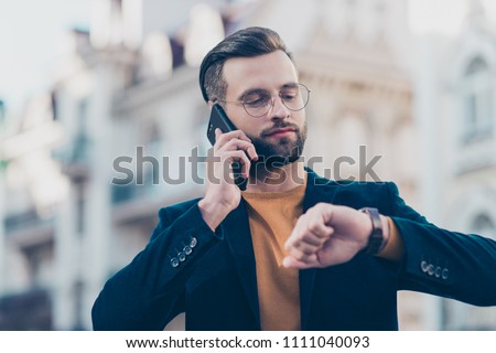 Hurry up! Portrait of responsible attractive man with modern hairdo having important call speaking by smart phone looking at luxury watch on wrist isolated on blurred background. Accessory concept Royalty-Free Stock Photo #1111040093