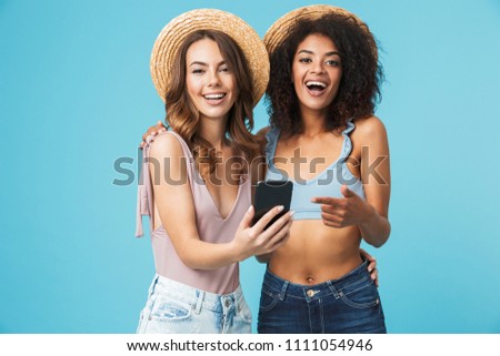 Happy photo of two multiethnic summer girls laughing and pointing finger at screen of smartphone while taking selfie isolated over blue background
