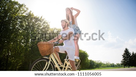 Low angle view of cheerful positive couple handsome man on bike beautiful woman standing on trunk watching meadow discovering new places enjoying time together. Active lifestyle