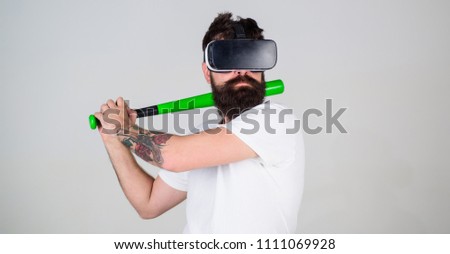 Man in VR glasses with hipster beard and tattoo posing with baseball bat isolated on gray background. Amateur player training batting skills in simulation game. Bearded man doing sports.