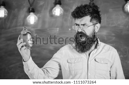 School bell concept. Bearded hipster holds clock, chalkboard on background, copy space. Teacher in eyeglasses holds alarm clock. Man with beard and mustache on angry shouting face looks at clock.