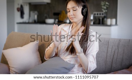 a young and pretty woman dressed in pajamas listening to music on headphones, a lady sitting in the living room on the couch, she sings and has fun with music, behind is the kitchen