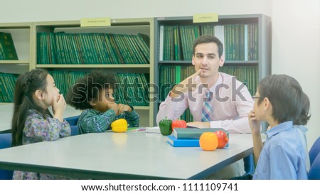 smiley caucasian teacher and grouping of asian kids student learning and talking at white table and color book with bookshelf background