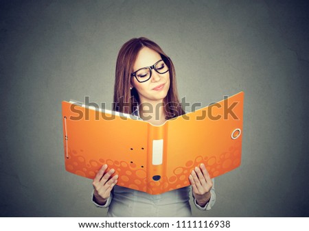 Young woman in glasses excited with education and reading huge folder with assignment on gray background 