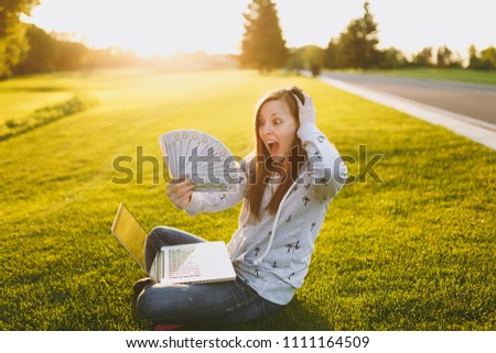 Female holding bundle of dollars cash money. Woman sitting on grass ground, working on laptop pc computer in city park on green grass sunshine lawn outdoors. Mobile Office. Freelance business concept