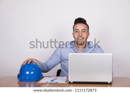 Young smiley employee sitting on his desk touches the blue helmet on the desk on a grey background.
