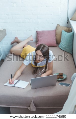 youth student with cookies using laptop and writing in copybook while lying on sofa at home 