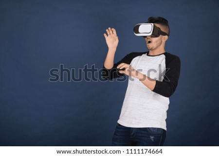 Young man standing, wearing the VR glasses raising his both hands frightened, on a blue background.
