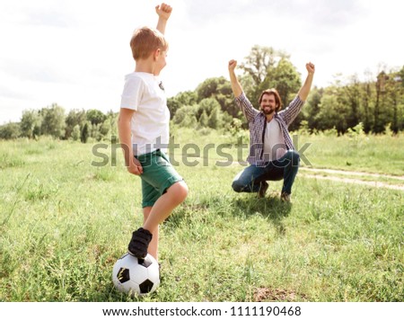 Father is screaming and yelling. He is very happy for his son. Boy played a good game. He won. Boy is standing besides his father and holding ball with leg.