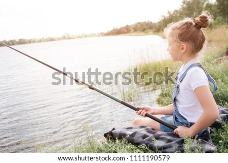 A picture of girl sitting alone at river shore. She is fishing. Girl is holding fish-rod with both hands. She is looking at water. Girl is very serious.