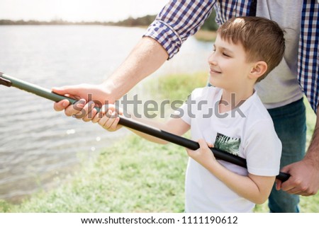 A picture of guy helping his son to hold fish-rod in a right way. He is holding it with one hand while boy is doing that with two hands. Small man seems like happy boy.