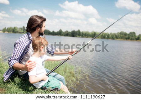 A picture of boys sitting together at the edge of lake and fishing. Boy is holding long fish-rod while his dad is guiding him to fishing right.