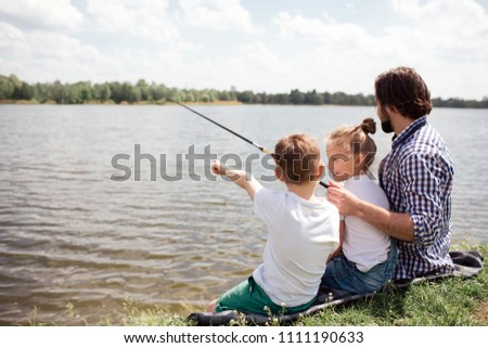 A picture of man and his children sitting together on the river shore. Guy is fishing while his kids are watching on it. Man is holding long fish-rod.