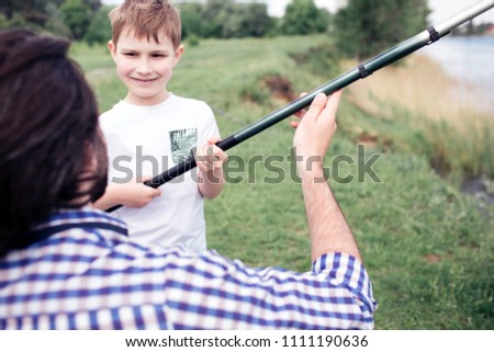 A picture of happy son standing in front of his dad and holding long fish-rod. Guy is holding it a bit. They are spending time together.