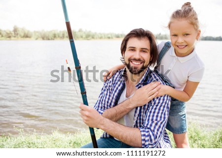 Man and girl are looking at camera and smiling. She is standing behind him and keeping her hands on his shoulders. Guy is holding fish-rod in left hand.