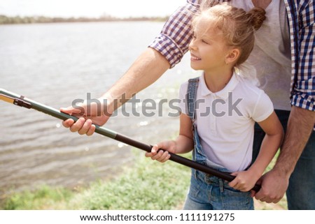 A picture of guy helping his daughter to hold fish-rod in a right way. Girl is holding it with both hands and smiling. She looks happy.